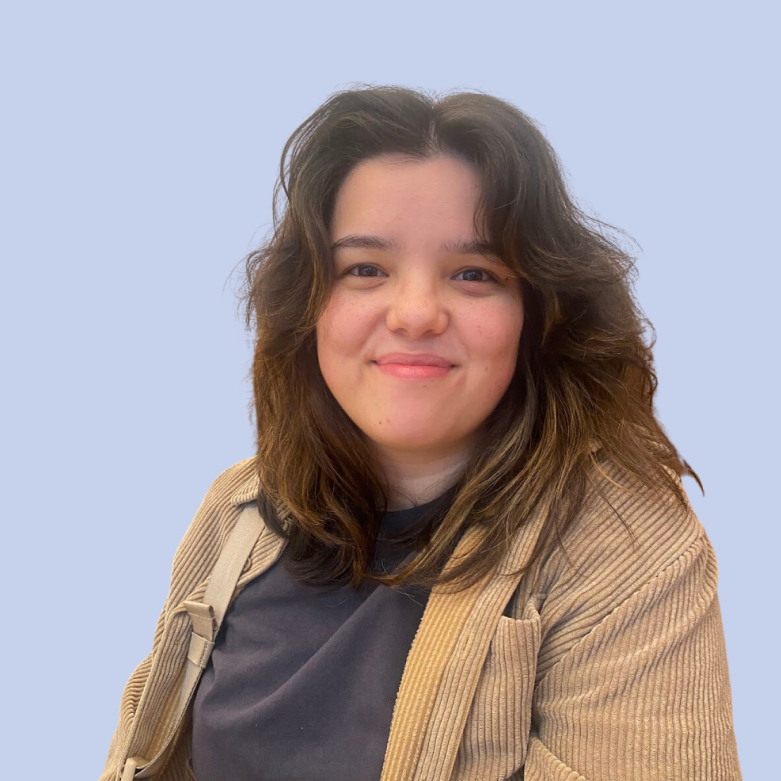 A person with shoulder-length dark hair, wearing a black shirt and beige jacket, smiles at the camera against a light blue background, appearing relaxed and approachable with a friendly expression, ideal for a social media planner | plannthat.com