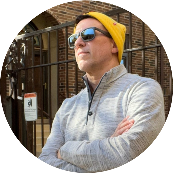 A man in a gray sweater and yellow beanie, wearing sunglasses, stands with arms crossed in front of a brick building with a gated entrance, looking confident and ready to use his social media scheduler | plannthat.com