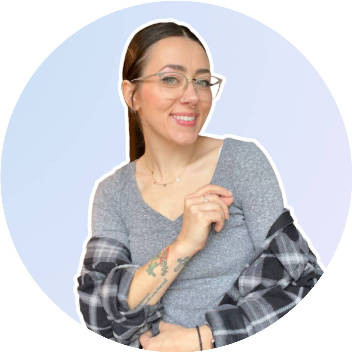A woman wearing glasses and a grey top, with a plaid shirt draped over her shoulders, smiles against a light blue background. She has tattoos on her right arm and wears a necklace, embodying the creative spirit of a social media planner | plannthat.com