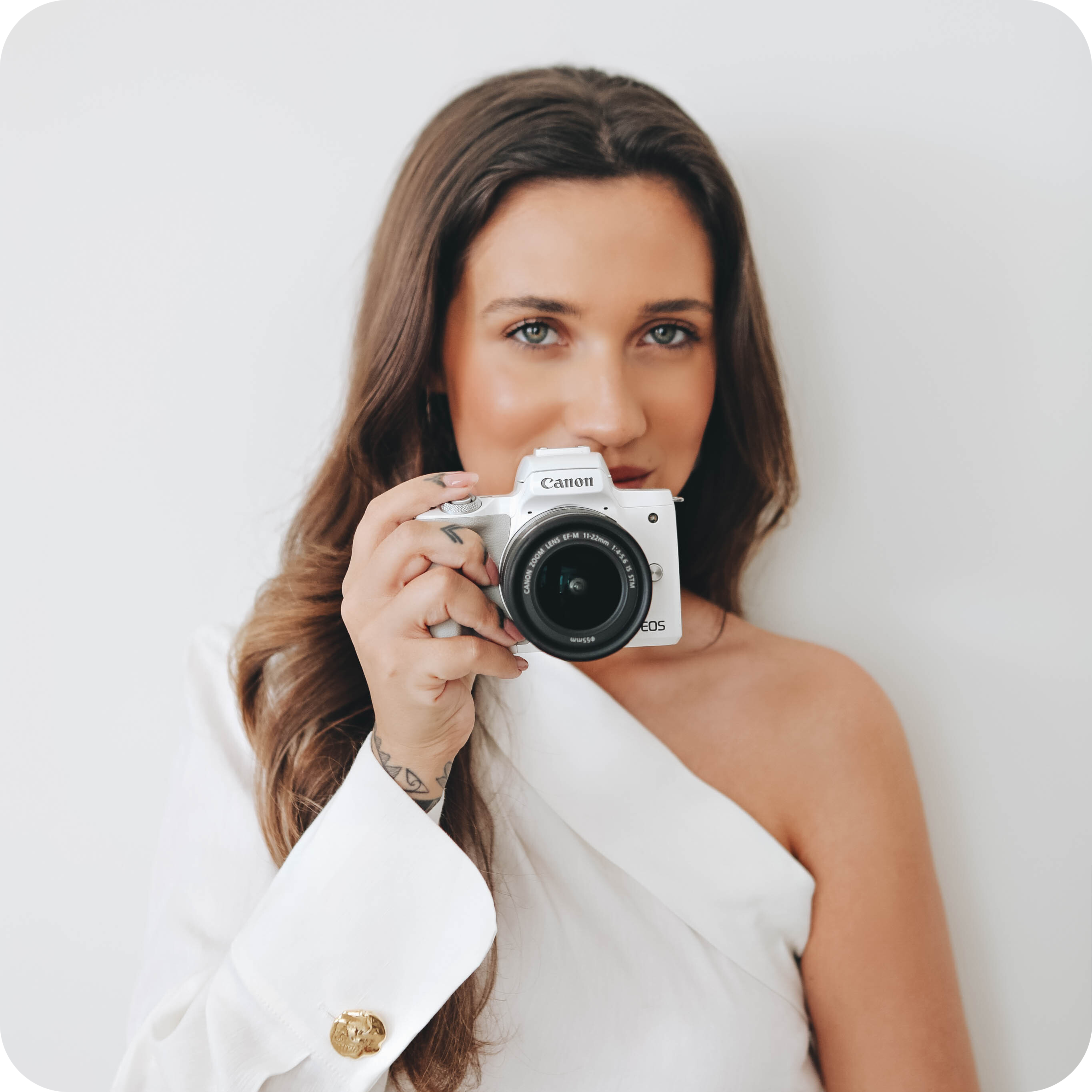 Woman in a white off-shoulder top holding a Canon EOS camera, looking directly at the lens, against a plain white background, perfect for a social media planner | plannthat.com